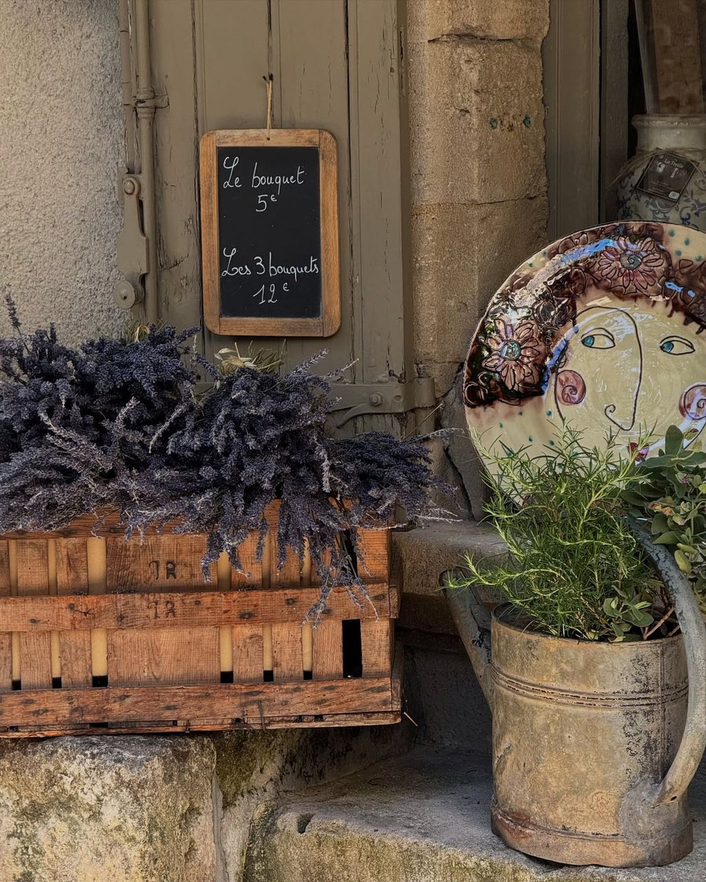 Decorative elements including a wooden crate with lavender, a rustic watering can with plants, and a chalkboard sign on a stone wall.
