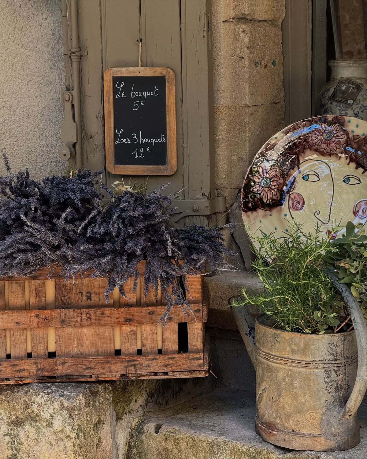 Decorative elements including a wooden crate with lavender, a rustic watering can with plants, and a chalkboard sign on a stone wall.