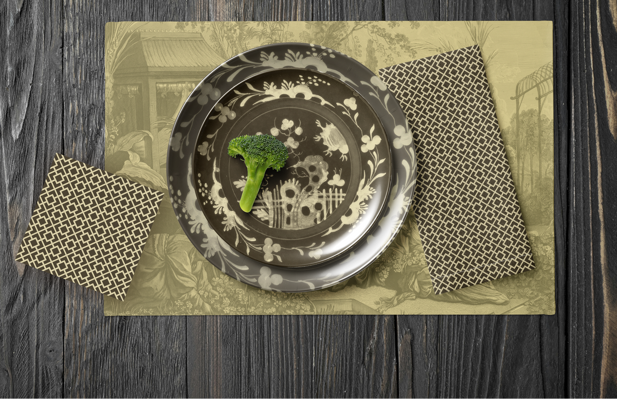 La Chinoiserie Fawn Party Table — plates stacked on the garden-scene placemat with fretwork napkins, in cream on umber, on dark wood