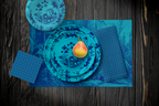 La Chinoiserie Lagoon Party Table — plates stacked on the garden-scene placemat with fretwork napkins, in navy on aqua, on dark wood
