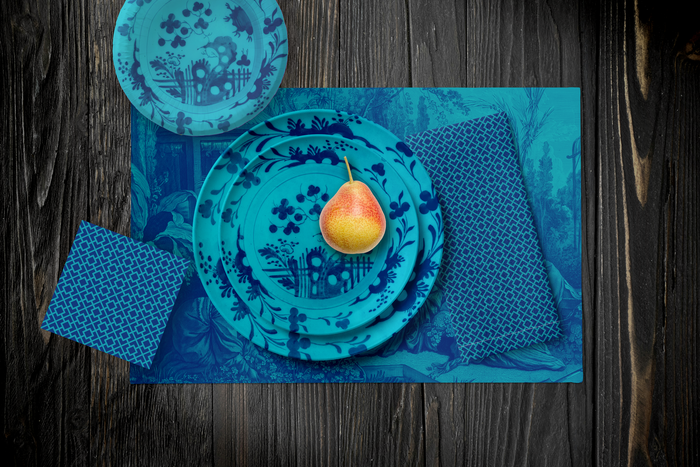 La Chinoiserie Lagoon Party Table — plates stacked on the garden-scene placemat with fretwork napkins, in navy on aqua, on dark wood