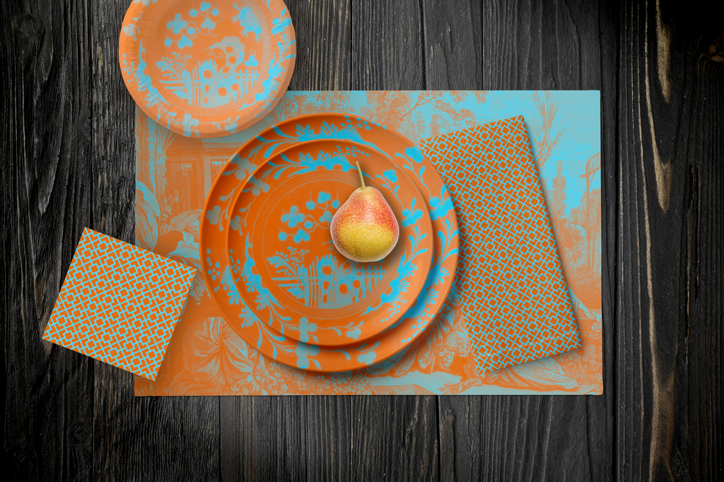 La Chinoiserie Orange Party Table — plates stacked on the garden-scene placemat with fretwork napkins, in aqua on orange, on dark wood