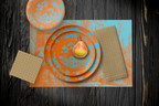 La Chinoiserie Orange Party Table — plates stacked on the garden-scene placemat with fretwork napkins, in aqua on orange, on dark wood