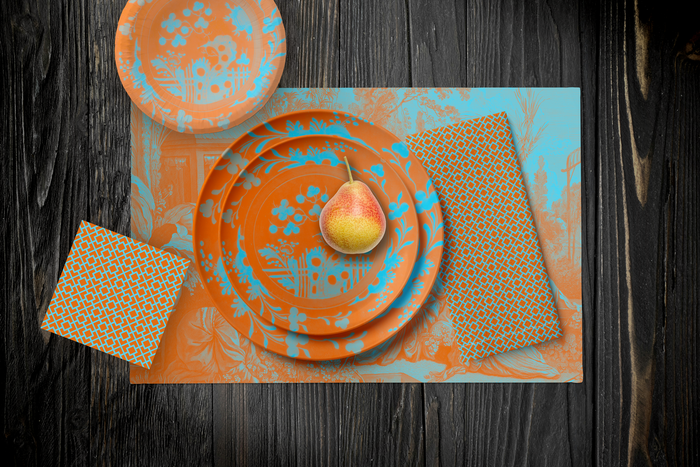 La Chinoiserie Orange Party Table — plates stacked on the garden-scene placemat with fretwork napkins, in aqua on orange, on dark wood