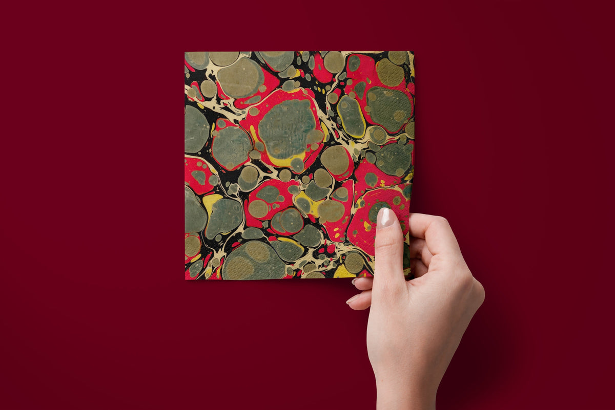 Noël Marbré Rouge paper napkins on a deep red ground — a napkin held in hand