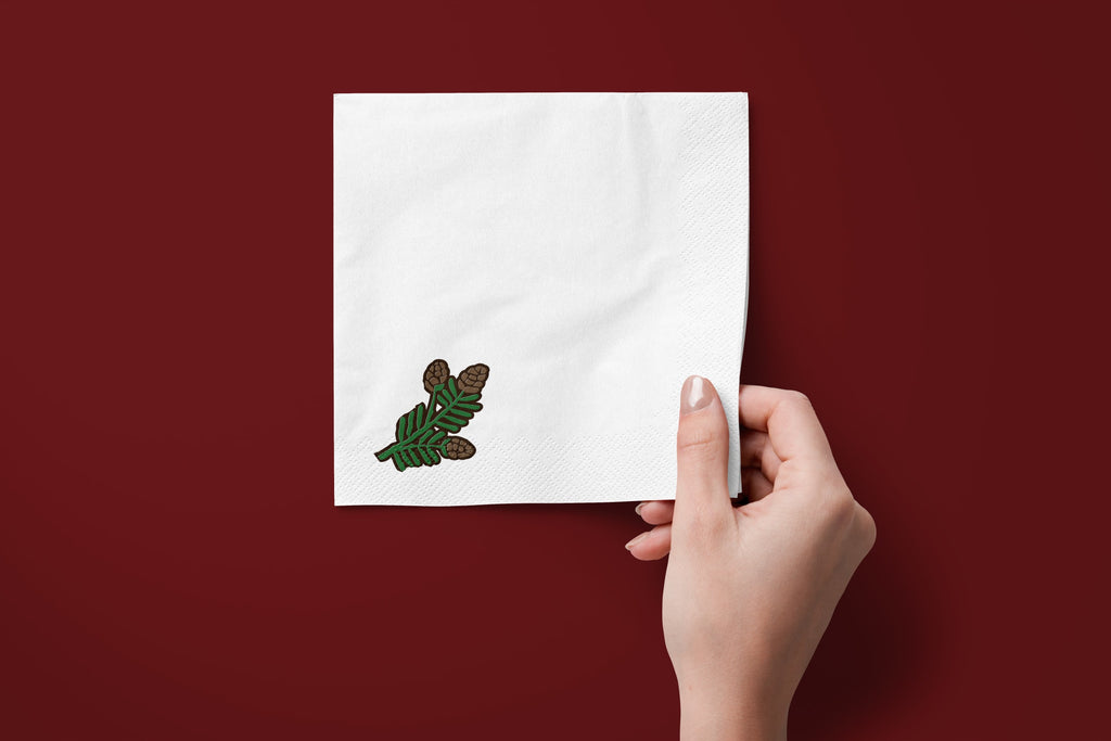 Noël Mini Pinecone paper napkins on a deep red ground — a napkin held in hand