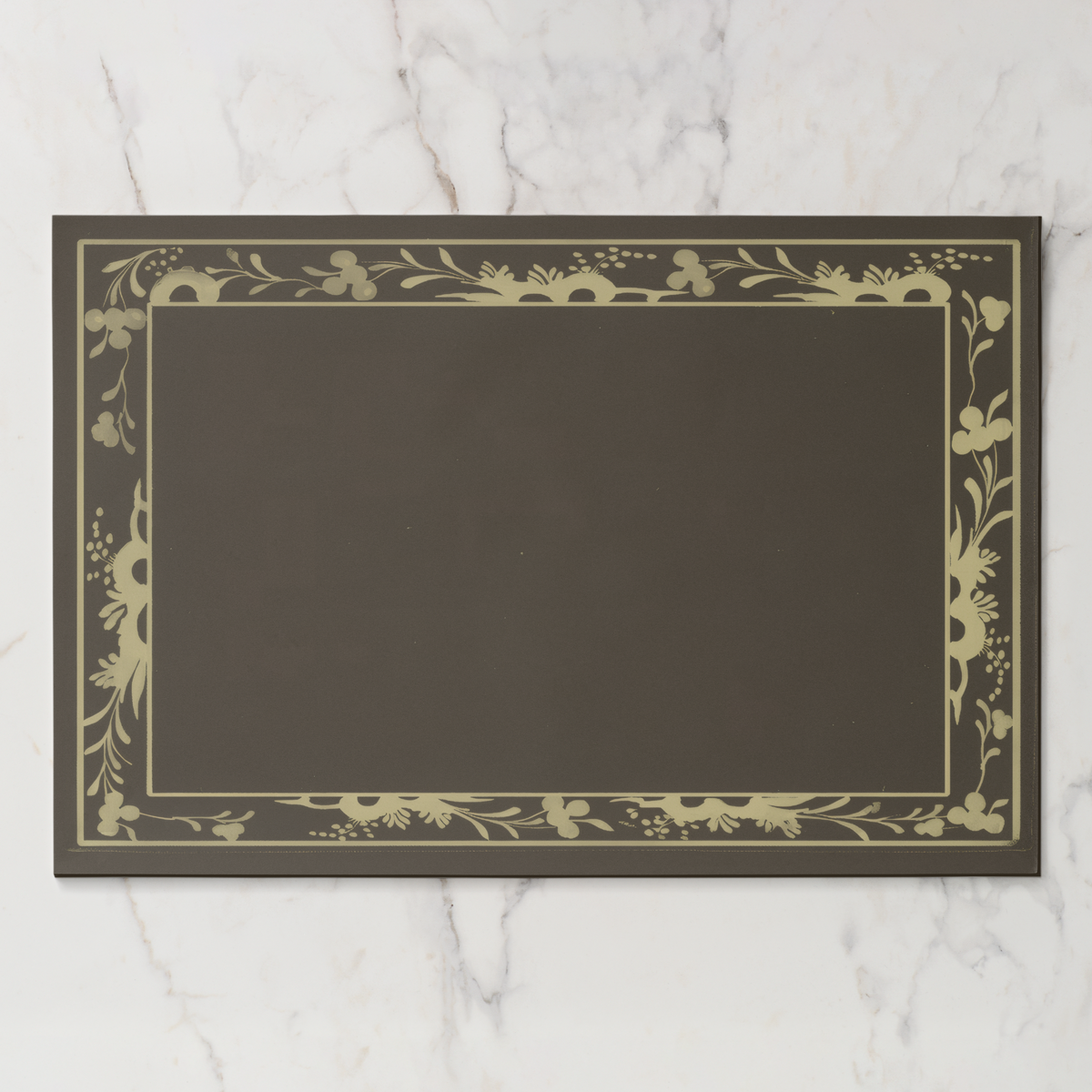 La Chinoiserie Bordure paper placemat in fawn — parchment engraved floral border on a deep umber ground