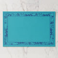 La Chinoiserie Bordure paper placemat in Lagoon — deep blue engraved floral border on turquoise, open centre