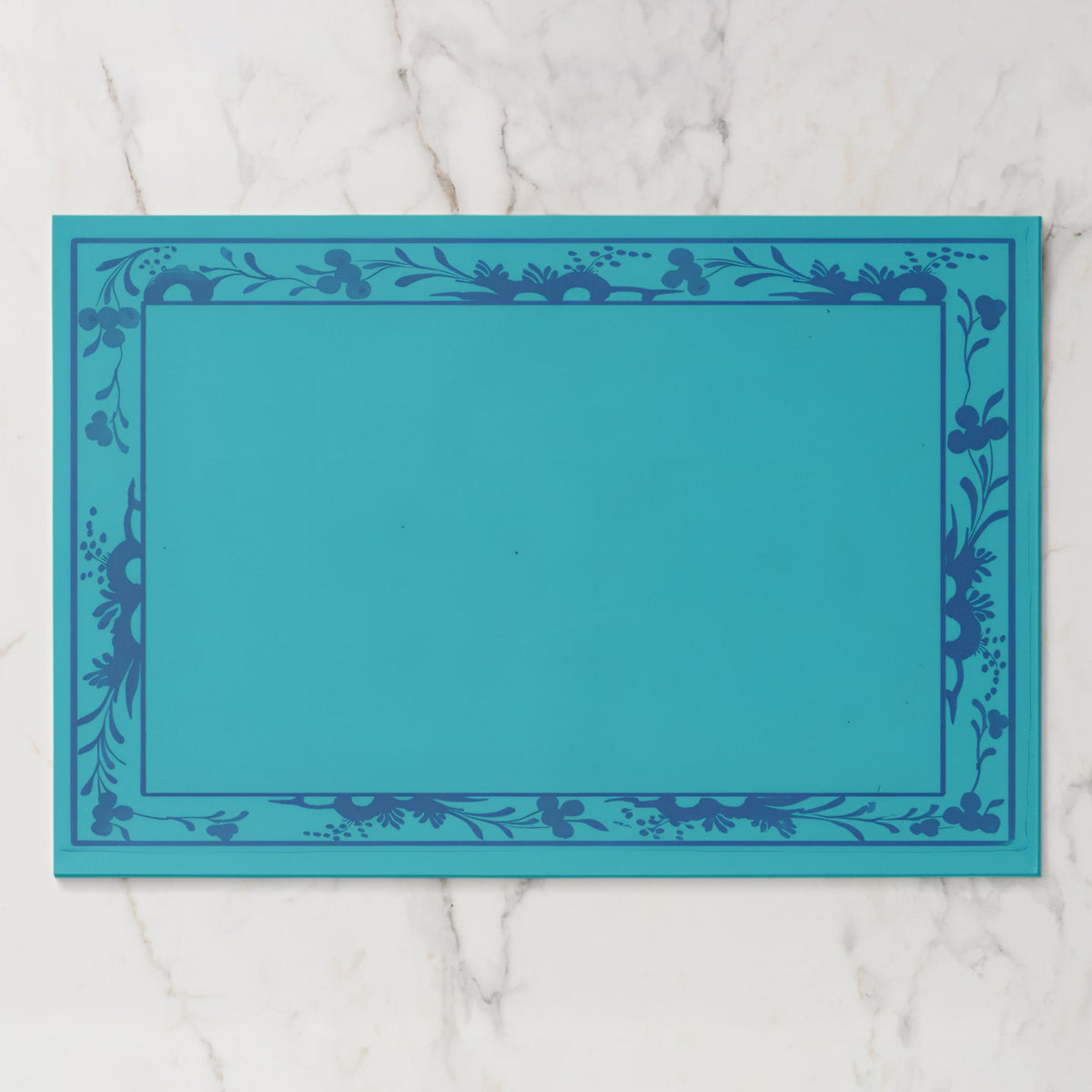 La Chinoiserie Bordure paper placemat in Lagoon — deep blue engraved floral border on turquoise, open centre