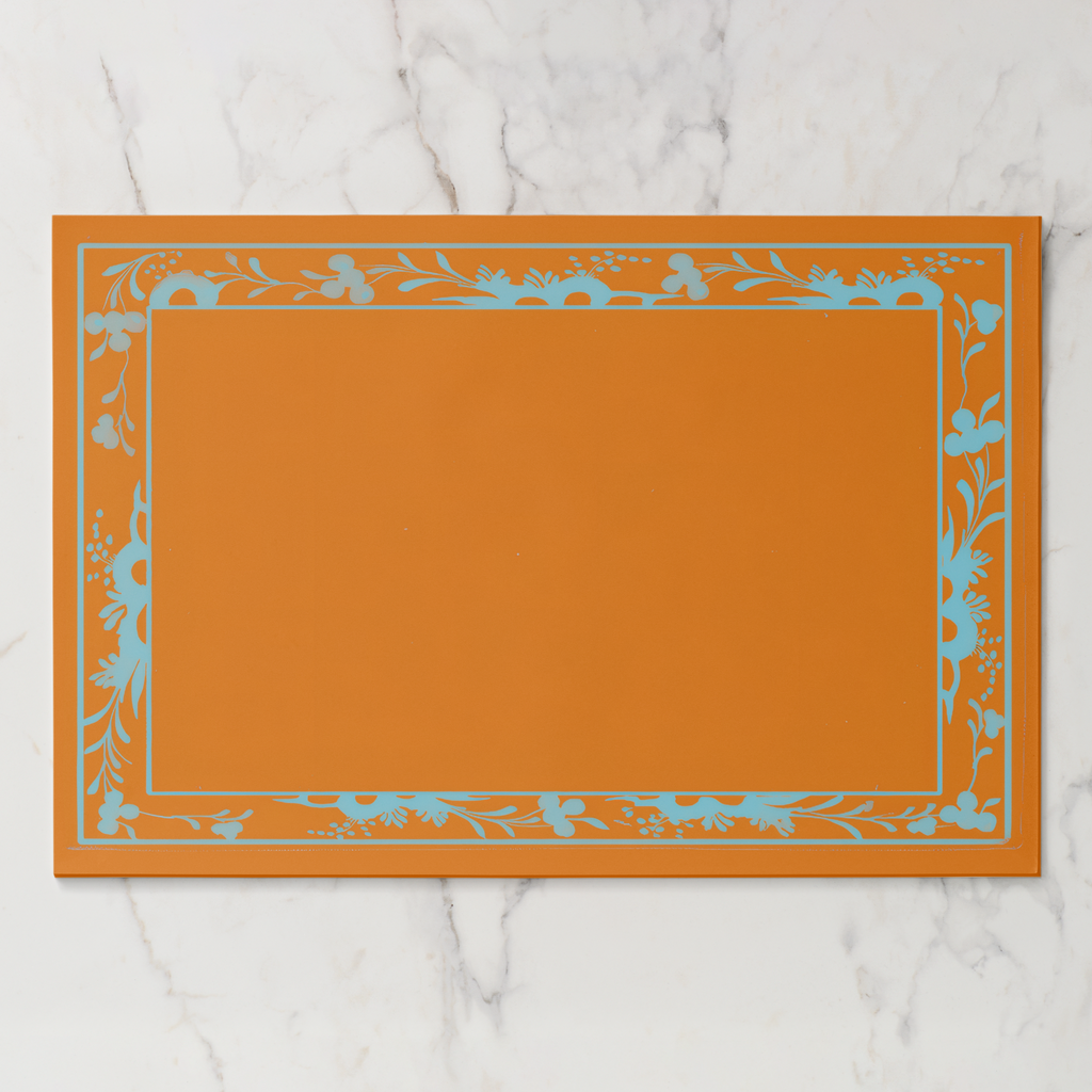 La Chinoiserie Bordure paper placemat in orange — pale blue engraved floral border on a tangerine ground