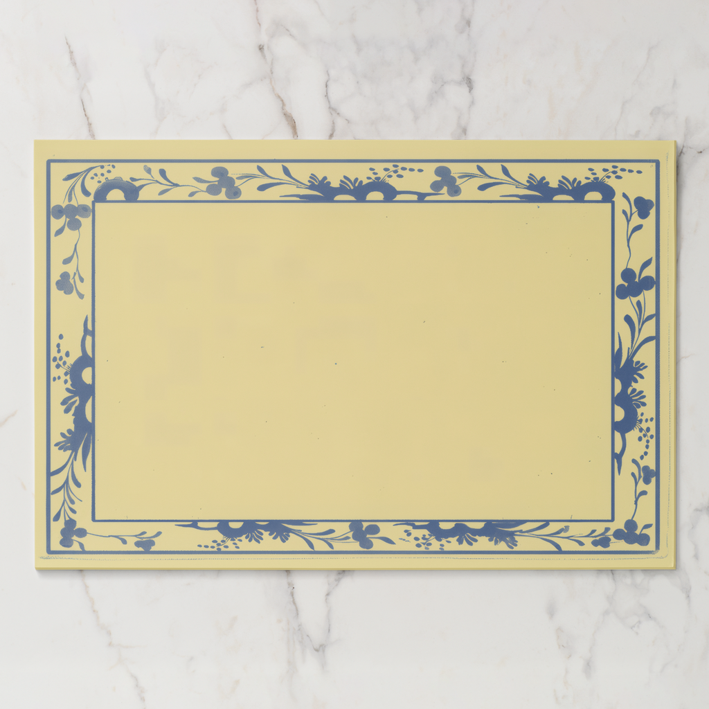 La Chinoiserie Bordure paper placemat in porcelain — navy engraved floral border on a butter-cream ground