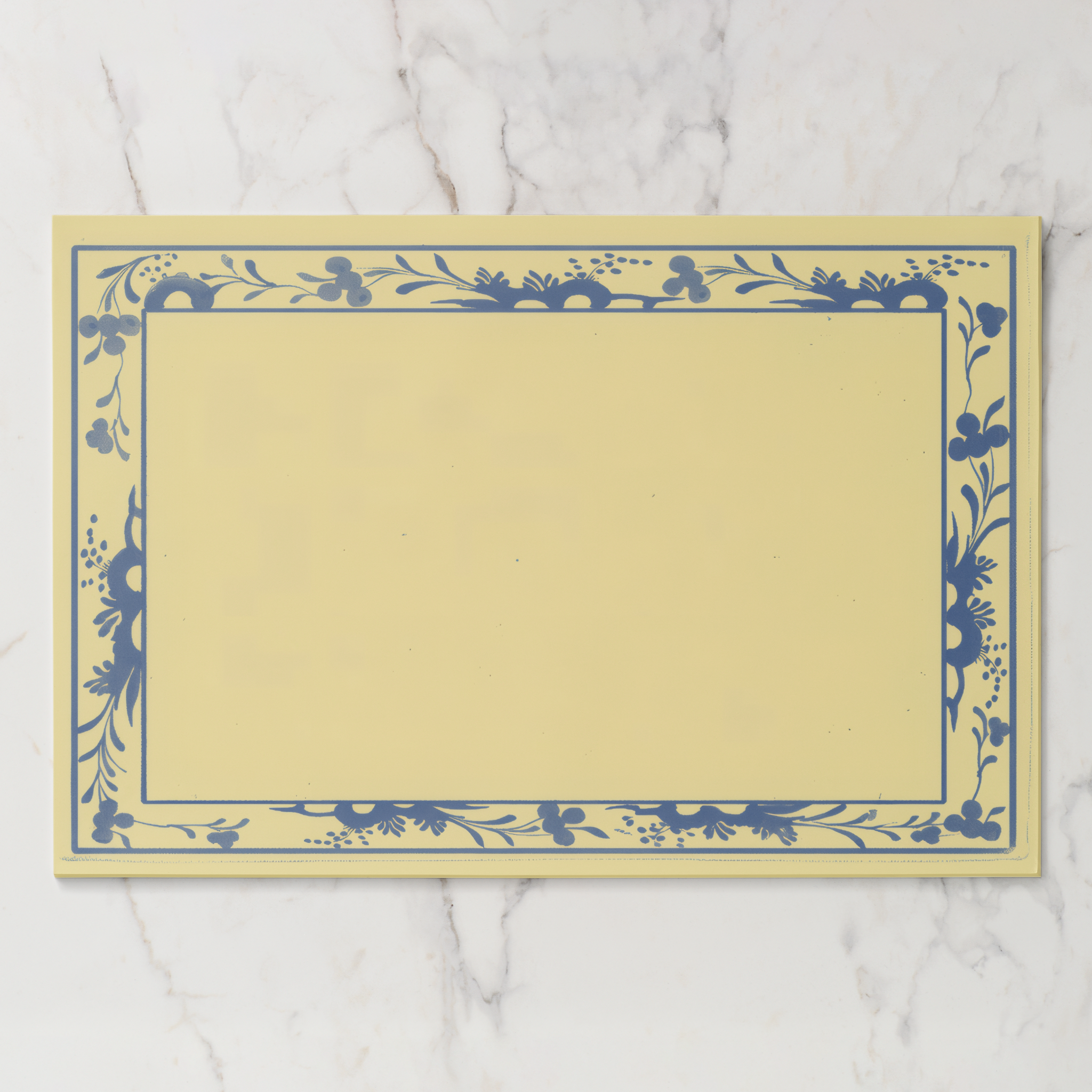 La Chinoiserie Bordure paper placemat in porcelain — navy engraved floral border on a butter-cream ground