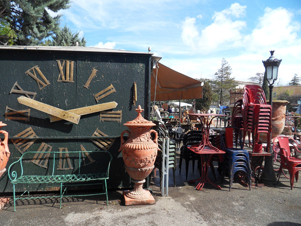 Vintage outdoor furniture and decor items including a large clock, urns, and chairs on a sunny day.